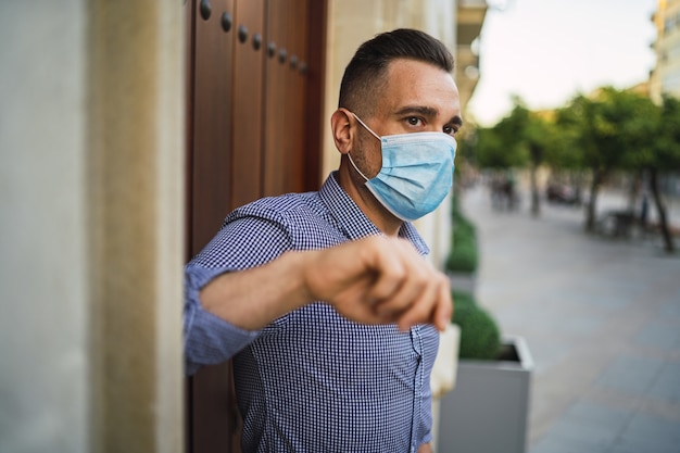 Junger Mann, der ein blaues Hemd trägt, das am Tor mit einer medizinischen Gesichtsmaske steht