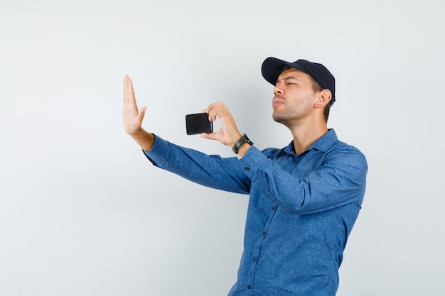 Junger Mann, der ein Foto von jemandem auf dem Handy in blauem Hemd, Mütze, Vorderansicht macht.