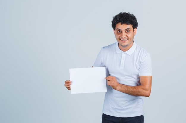 Junger Mann, der ein leeres Papierblatt im weißen T-Shirt, in der Hose hält und selbstbewusst aussieht, Vorderansicht.