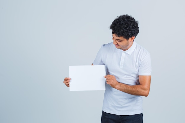 Junger Mann, der ein leeres Papierblatt im weißen T-Shirt, in der Hose hält und selbstbewusst aussieht, Vorderansicht.