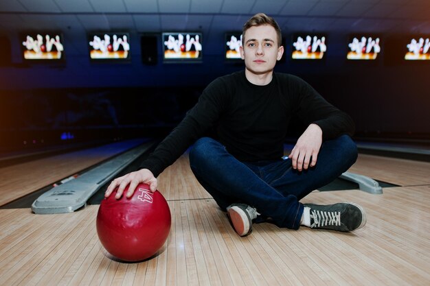 Junger Mann, der eine Bowlingkugel hält, die mit ultraviolettem Licht gegen Bowlingbahnen sitzt