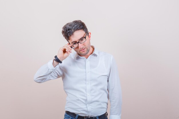 Junger Mann, der eine Brille in weißem Hemd, Jeans und nachdenklichem Blick abnimmt