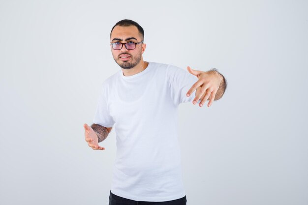 Junger Mann, der eine Brille trägt und sich die Hände ausstreckt, als er etwas in weißem T-Shirt und schwarzer Hose hält und glücklich aussieht?