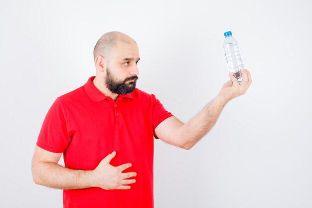 Junger Mann, der eine Flasche Wasser im roten Hemd, Vorderansicht betrachtet.