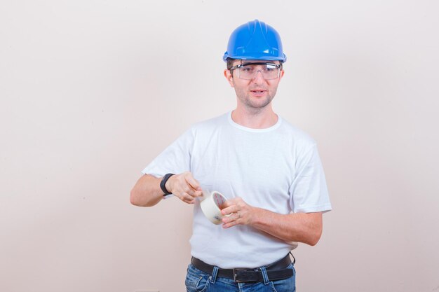 Junger Mann, der eine Rolle Klebeband in T-Shirt, Jeans, Helm öffnet und vorsichtig aussieht