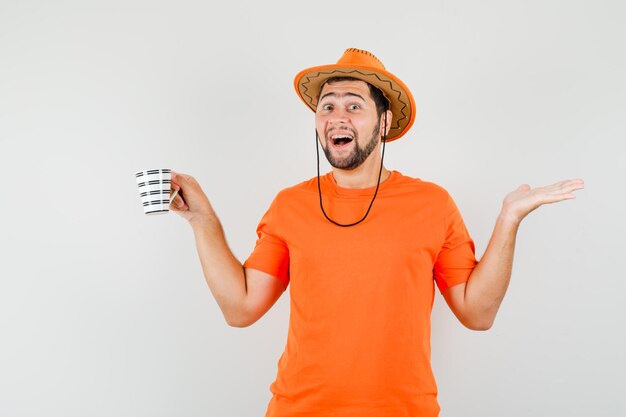 Junger Mann, der eine Tasse Getränk in orangefarbenem T-Shirt, Hut und fröhlichem Aussehen hält. Vorderansicht.