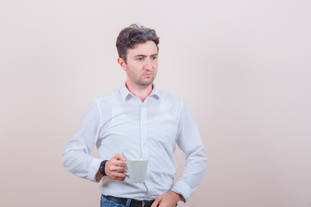 Junger Mann, der eine Tasse Getränk in weißem Hemd, Jeans und nachdenklichem Blick hält