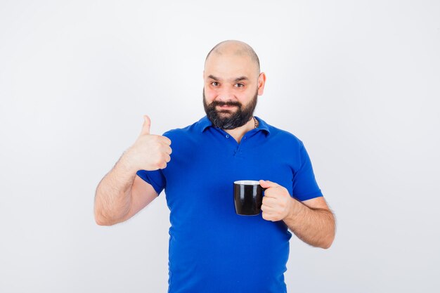 Junger Mann, der eine Tasse hält, während er den Daumen im blauen Hemd zeigt und zufrieden aussieht, Vorderansicht.