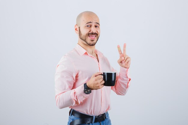 Junger Mann, der eine Tasse hält, während er die Siegesgeste in rosa Hemd, Jeans zeigt und fröhlich aussieht. Vorderansicht.