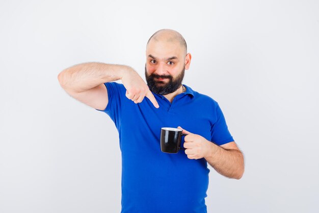 Junger Mann, der eine Tasse hält, während er im blauen Hemd darauf zeigt und optimistisch aussieht. Vorderansicht.