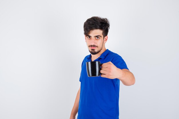 Junger Mann, der eine Tasse im blauen T-Shirt hält und ernst schaut