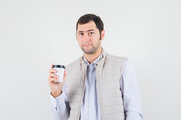 Junger Mann, der eine Tasse Kaffee in beiger Jacke und Kappe hält und ernst schaut. Vorderansicht.