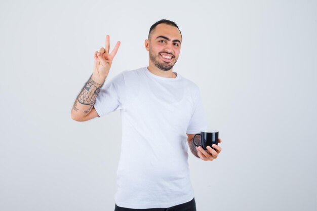 Junger Mann, der eine Tasse Tee hält und ein Friedenszeichen in weißem T-Shirt und schwarzer Hose zeigt und glücklich aussieht