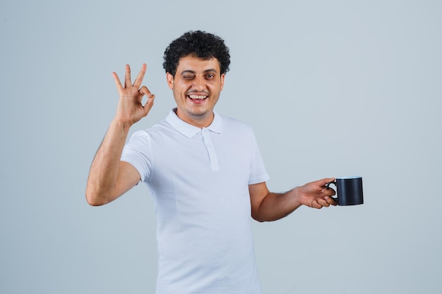Junger Mann, der eine Tasse Tee hält, während er ein OK-Zeichen zeigt und in weißem T-Shirt und Jeans zwinkert und glücklich aussieht. Vorderansicht.