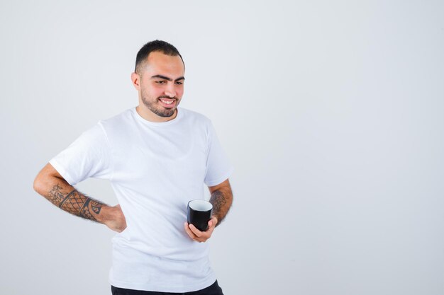 Junger Mann, der eine Tasse Tee in einer Hand hält und die Hand in einem weißen T-Shirt und einer schwarzen Hose an der Taille hält und glücklich aussieht