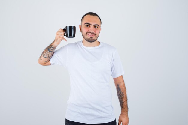 Junger Mann, der eine Tasse Tee in weißem T-Shirt und schwarzer Hose hält und glücklich aussieht