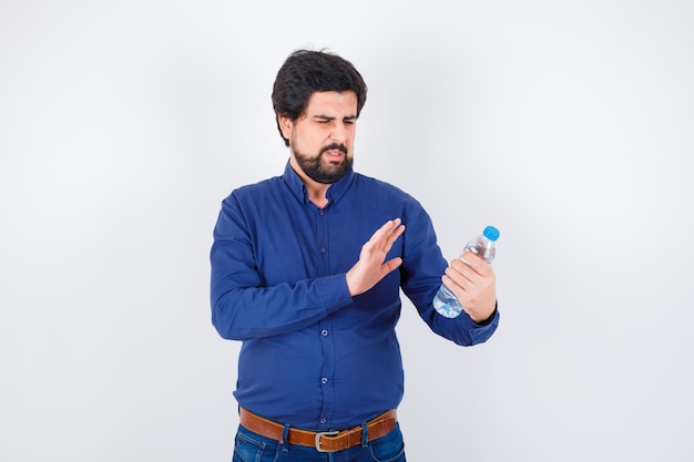 Junger Mann, der eine Wasserflasche hält und die Hand in blauem Hemd und Jeans darauf ausstreckt und ernst aussieht, Vorderansicht.