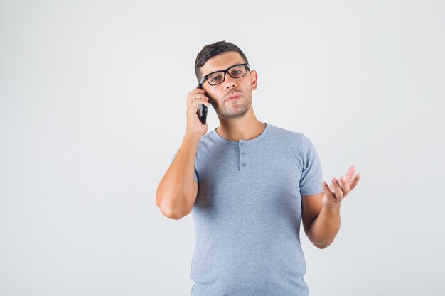 Junger Mann, der einen Anruf in grauem T-Shirt, Brille macht und nachdenklich aussieht