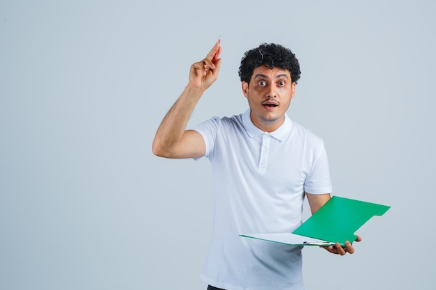 Junger Mann, der einen Stift in der Heureka-Geste anhebt und ein Notizbuch in weißem T-Shirt und Jeans hält und vernünftig aussieht. Vorderansicht.