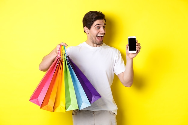 Junger Mann, der Einkaufstaschen hält und Handybildschirm, Geldanwendung zeigt, über gelbem Hintergrund stehend.
