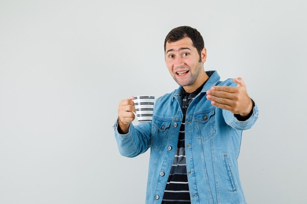 Junger Mann, der einlädt, zu kommen, während Tasse des Getränks im T-Shirt, in der Jacke hält und lustig schaut