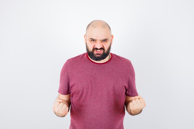 Junger Mann, der Erfolgsgeste in rosa T-Shirt zeigt und mächtig aussieht, Vorderansicht.