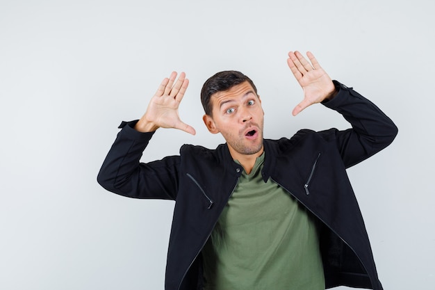 Junger mann, der erhobene handflächen in der luft in t-shirt, jacke hält und amüsiert aussieht. vorderansicht.