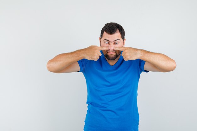 Junger Mann, der Finger auf Nase in blauem T-Shirt drückt und seltsam aussieht