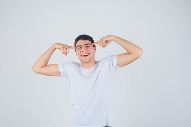 Junger Mann, der Finger auf Schläfen im T-Shirt hält und glücklich schaut. Vorderansicht.