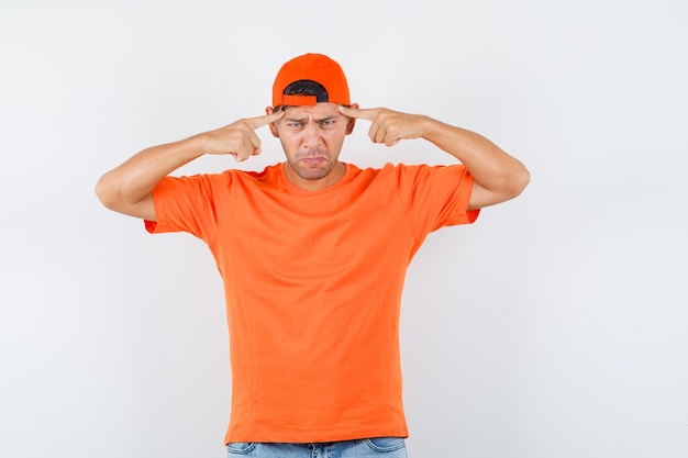 Junger Mann, der Finger auf Schläfen in orange T-Shirt und Mütze, Jeans hält und schmerzhaft aussieht