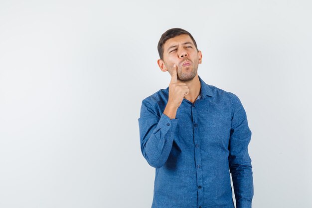 Junger Mann, der Finger auf Wange im blauen Hemd hält und nachdenklich schaut, Vorderansicht.