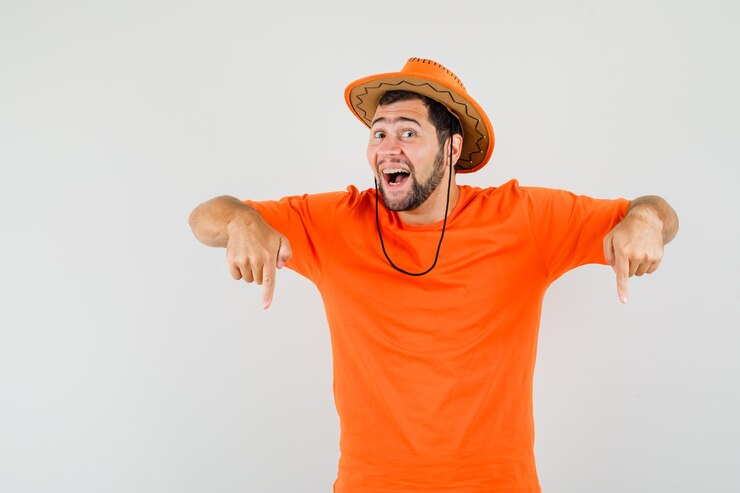 Kostenlose Foto junger mann, der finger unten in orange t-shirt, hut zeigt und freudig schaut, vorderansicht.