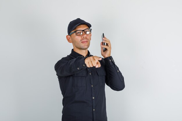 Junger Mann, der Finger zur Kamera mit Smartphone im schwarzen Hemd mit Kappe zeigt