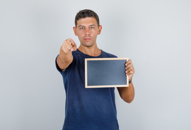 Junger Mann, der Finger zur Kamera mit Tafel im dunkelblauen T-Shirt, Vorderansicht zeigt.