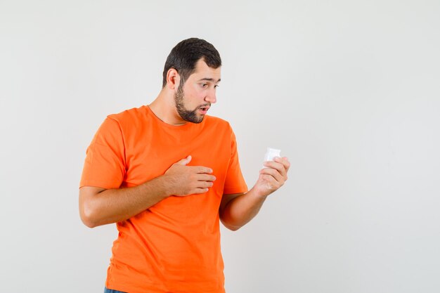 Junger Mann, der Flasche der Pillen im orangefarbenen T-Shirt betrachtet und verwirrt schaut. Vorderansicht.