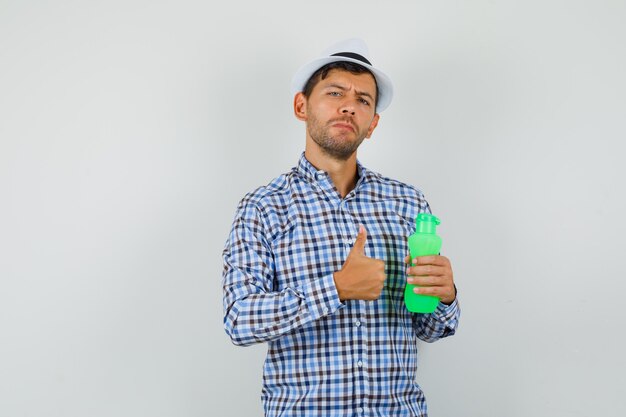 Junger Mann, der Flasche für Wasser hält und Daumen oben im karierten Hemd zeigt