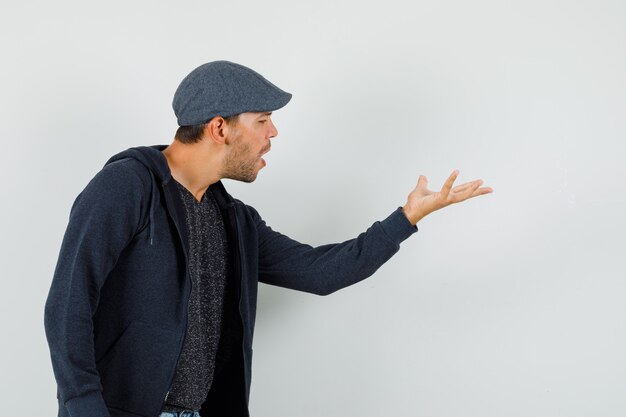 Junger Mann, der fragende Geste in T-Shirt, Jacke, Mütze macht und nervös schaut.