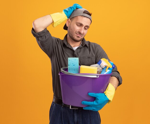 Junger Mann, der Freizeitkleidung und Mütze in Gummihandschuhen hält, die Eimer mit Reinigungswerkzeugen halten, verwirrt und unzufrieden mit der Hand auf seinem Kopf stehend über orange Wand