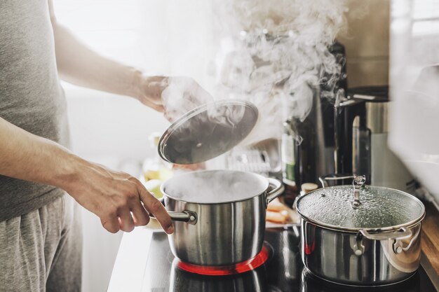 Junger Mann, der frisches Essen zu Hause kocht und Deckel des dampfenden Topfes öffnet.