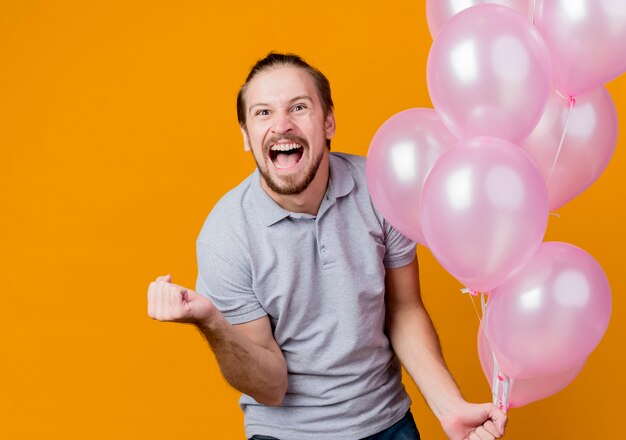 Junger mann, der geburtstagsfeier feiert, die bündel der ballonschlifffaust verrückt glücklich und aufgeregt schreit, die über orange wand stehen stehen