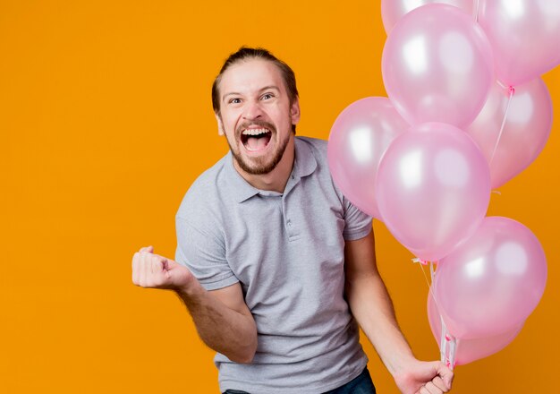 Junger Mann, der Geburtstagsfeier feiert, die Bündel der Ballonschlifffaust verrückt glücklich und aufgeregt schreit, die über orange Wand stehen stehen