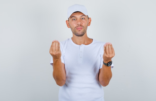 Junger Mann, der Geldgeste mit Händen im weißen T-Shirt, Kappe, Vorderansicht tut.