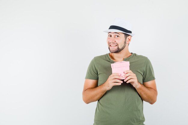 Junger Mann, der Geschenkbox im grünen T-Shirt und im Hut hält und niedlich schaut