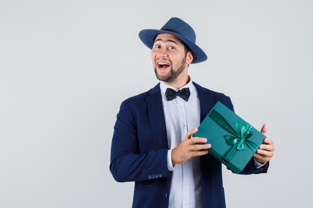 Junger Mann, der Geschenkbox in Anzug, Hut hält und glücklich schaut. Vorderansicht.