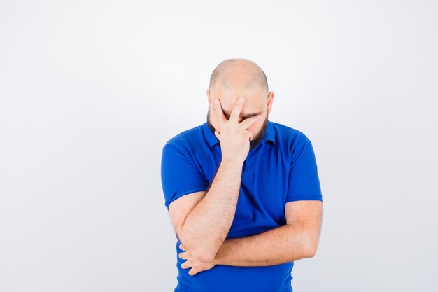 Junger Mann, der Gesicht mit der Hand im blauen Hemd bedeckt und verärgert schaut. Vorderansicht.