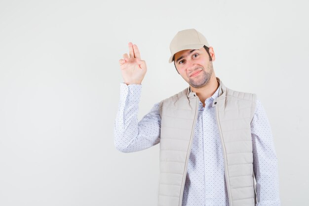 Junger Mann, der Gewehrzeichen in beige Jacke und Kappe zeigt und glückliche Vorderansicht schaut.