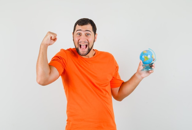 Junger Mann, der Globus mit Gewinnergeste im orangefarbenen T-Shirt hält und glücklich aussieht. Vorderansicht.