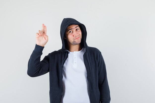 Junger Mann, der Grußgeste in T-Shirt, Jacke zeigt und selbstbewusst aussieht. Vorderansicht.