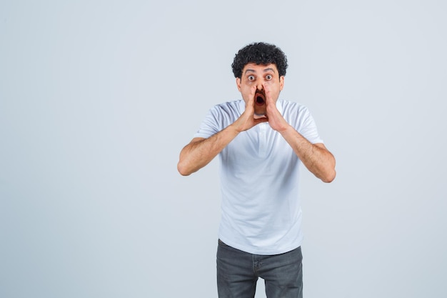 Junger Mann, der Händchen in der Nähe des Mundes hält, als er jemanden in weißem T-Shirt und Jeans anruft und überrascht aussieht, Vorderansicht.