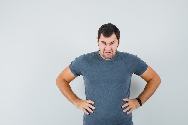 Junger Mann, der Hände auf Taille im grauen T-Shirt hält und wütend schaut